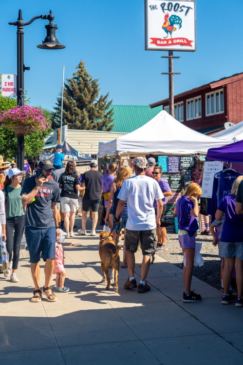 A busy street market scene with people walking, tents set up, and a dog on a leash under a clear blue sky.