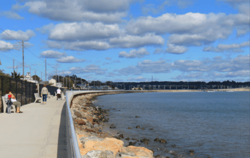 A scenic waterfront path with people walking, rocky shore, and a blue sky with clouds in the background.