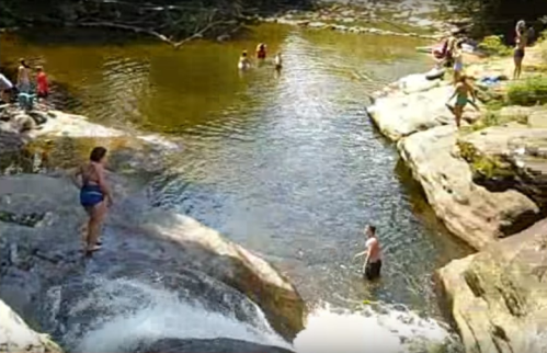 People enjoying a sunny day by a natural swimming hole with rocks and water. Some are swimming, others relaxing.