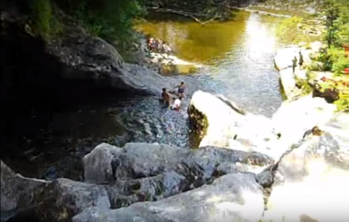 A scenic view of a rocky riverbank with people enjoying the water and lush greenery surrounding the area.