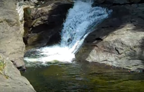 A small waterfall cascades over rocky terrain into a clear pool below, surrounded by natural greenery.