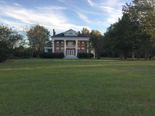 A large, two-story brick house with white columns, surrounded by green grass and trees under a blue sky.
