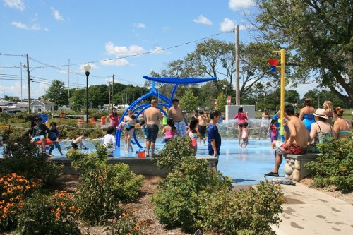 A lively splash pad filled with children and adults enjoying water play on a sunny day, surrounded by greenery and flowers.