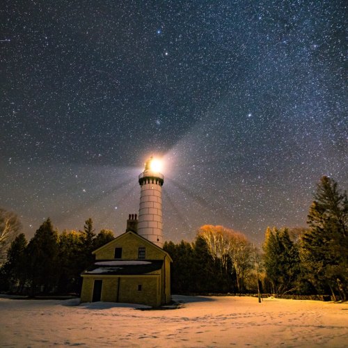 A lighthouse shines brightly under a starry night sky, surrounded by trees and snow-covered ground.