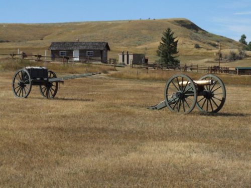 Two old wooden wagons sit in a grassy field, with a rustic cabin and hills in the background under a clear blue sky.