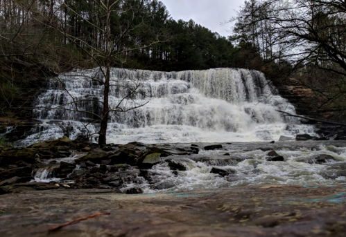 A cascading waterfall flows over rocks, surrounded by trees and a cloudy sky.