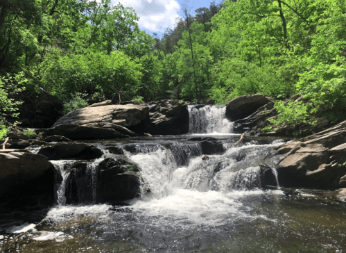 A serene waterfall cascades over rocks, surrounded by lush green trees and a bright blue sky.