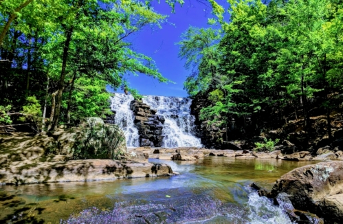 A serene waterfall cascades over rocks, surrounded by lush green trees under a bright blue sky.