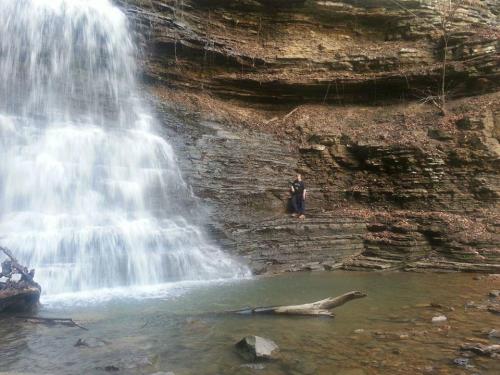 A serene waterfall cascades over rocky cliffs, with a person sitting peacefully by the water's edge.