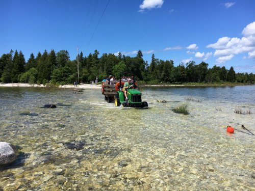 A tractor wades through shallow water, surrounded by trees and a clear blue sky, with people in the background.