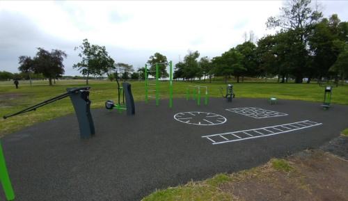 Outdoor fitness area with green equipment on a rubber surface, surrounded by trees and open grass.