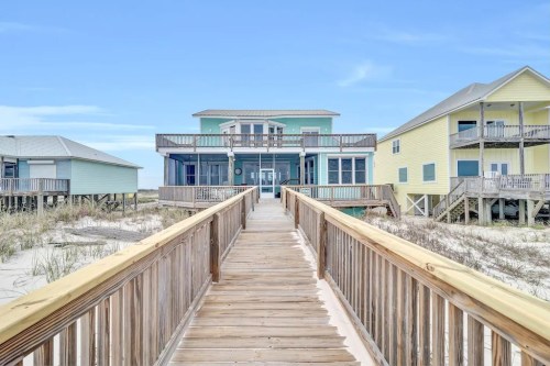 A wooden walkway leads to a beach house, flanked by two colorful houses under a clear blue sky.