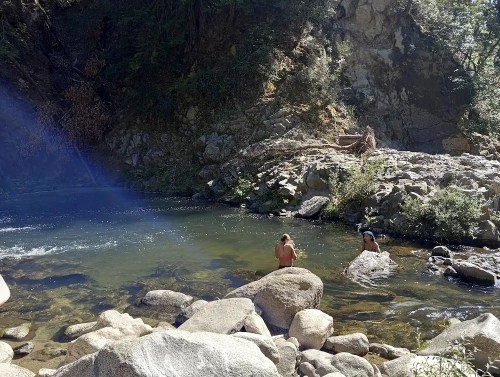 Two people wade in a clear river surrounded by rocky terrain and lush greenery. Sunlight filters through the trees.