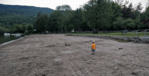 A young child in an orange shirt stands on a sandy beach near a calm lake, surrounded by trees and mountains.