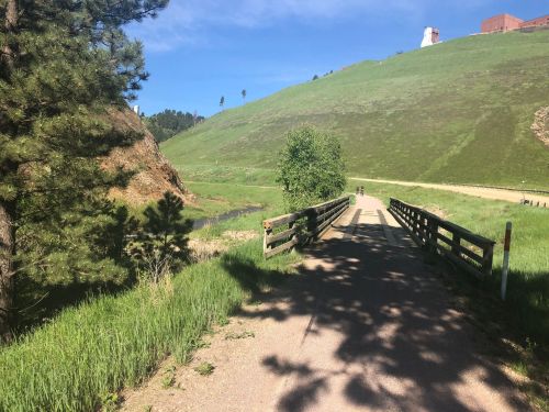 A scenic pathway with a wooden bridge, surrounded by green hills and trees under a clear blue sky.