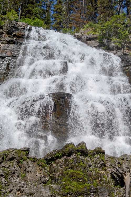 A cascading waterfall flows down rocky cliffs, surrounded by lush green trees under a clear blue sky.