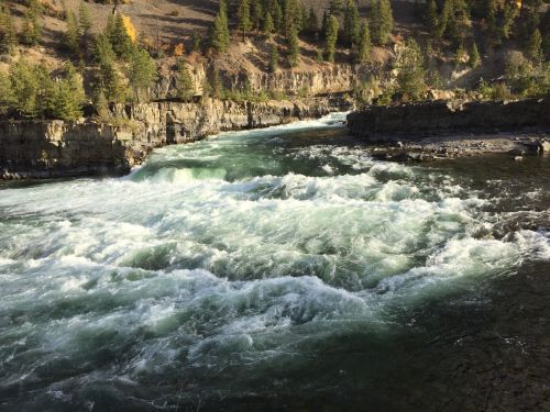 A rushing river flows through rocky terrain, surrounded by trees and a steep, rugged landscape.