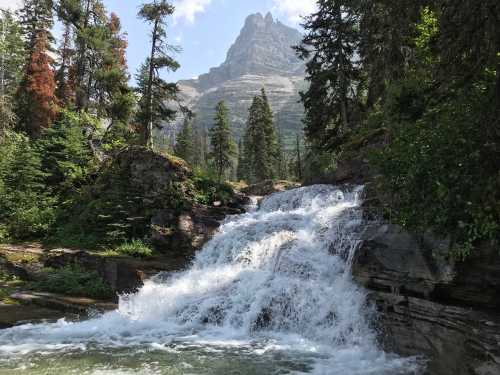 A cascading waterfall flows over rocks, surrounded by lush greenery and towering mountains under a clear blue sky.