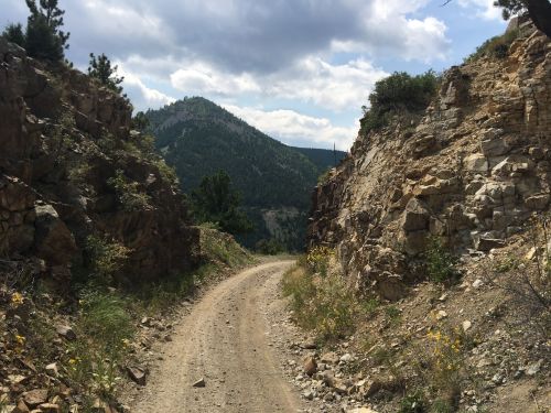 A dirt path winds between rocky cliffs, leading into a lush green valley under a cloudy sky.