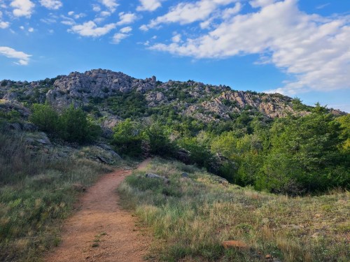 A winding dirt path leads through green grass and shrubs towards a rocky hill under a blue sky with scattered clouds.