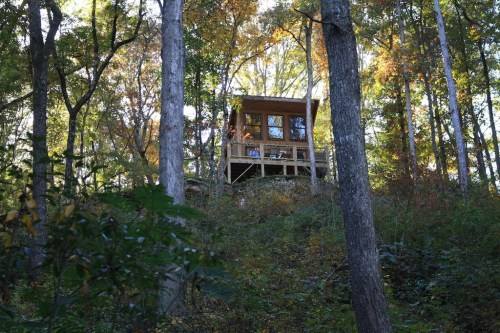 A wooden cabin perched on a hillside, surrounded by trees with autumn foliage.