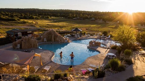 A scenic pool area at sunset, featuring rocky formations, lounge chairs, and two people enjoying the water.