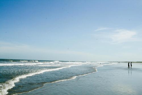Two people walk along a sandy beach with gentle waves under a clear blue sky.