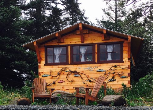 A cozy wooden cabin with large windows, surrounded by greenery and two Adirondack chairs in front.