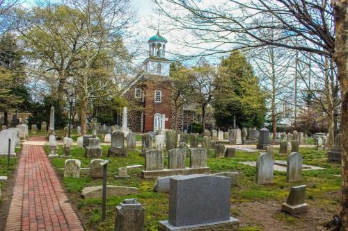 A peaceful cemetery with gravestones, a brick path, and a historic building in the background under a cloudy sky.