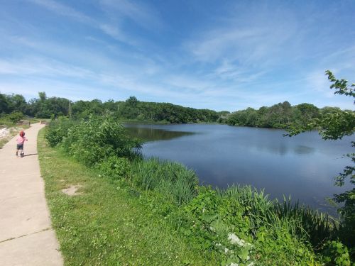 A child walks along a path beside a calm lake, surrounded by lush greenery and a clear blue sky.