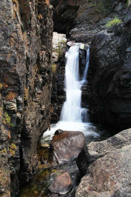 A serene waterfall cascades between rocky cliffs, surrounded by lush greenery and smooth stones.