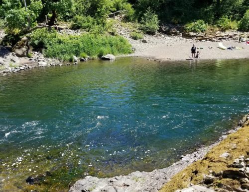 A serene river scene with clear water, a sandy shore, and people enjoying the outdoors among lush greenery.