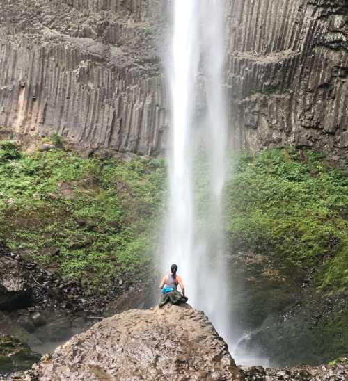 A person sits on a rock in front of a tall waterfall, surrounded by lush greenery and rocky cliffs.