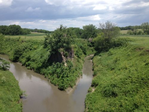 A winding river flows through lush green banks under a cloudy sky, surrounded by trees and fields.