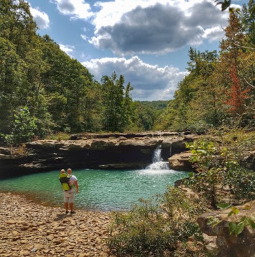 A person stands by a serene, green pool with a waterfall, surrounded by lush trees and a cloudy sky.