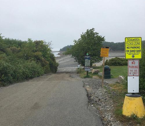 A gravel road leads to a beach area with signs indicating no parking and flood zone warnings. Overcast sky in the background.