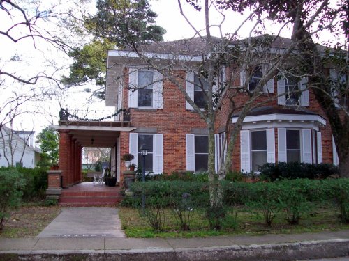 A two-story brick house with white shutters, surrounded by trees and shrubs, featuring a front porch and walkway.