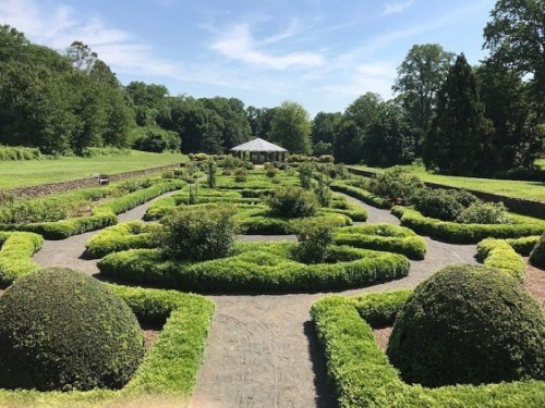 A lush garden with neatly trimmed hedges, circular topiaries, and a gazebo in the background under a clear blue sky.