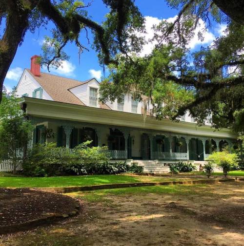 A historic house with a wraparound porch, surrounded by lush greenery and large trees under a bright blue sky.