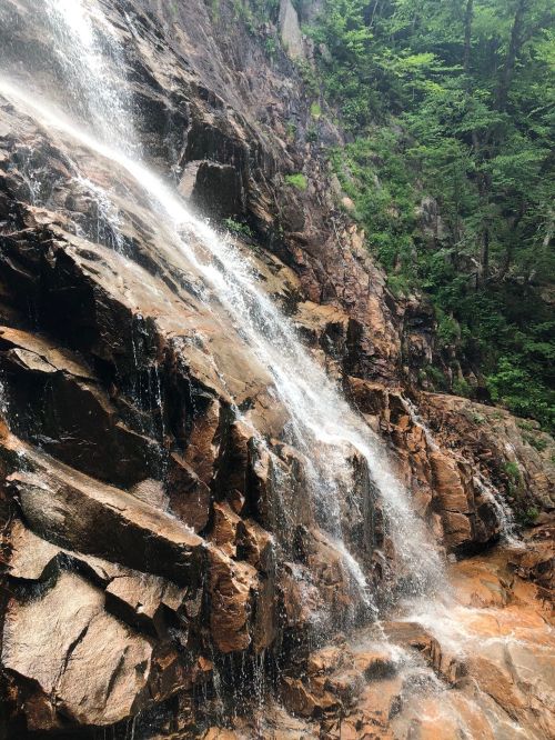A cascading waterfall flows over rocky terrain, surrounded by lush green foliage and steep cliffs.
