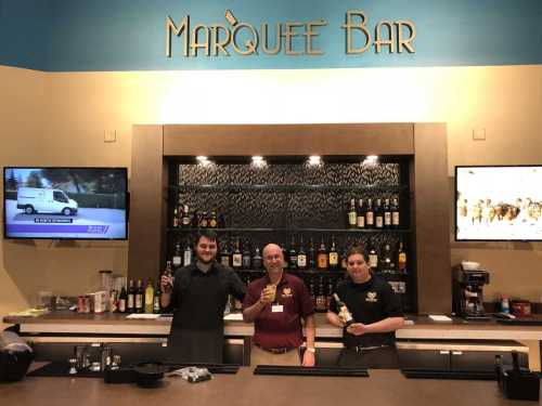 Three bartenders stand behind a bar, smiling and holding drinks, with shelves of liquor and screens in the background.