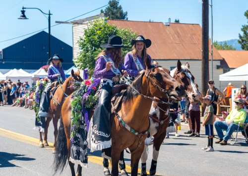 Three women in purple outfits ride horses adorned with flowers during a parade, with a crowd watching in the background.
