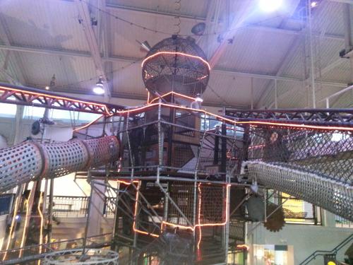 A large indoor play structure with nets, tubes, and lights, resembling a tower, in a spacious recreational area.