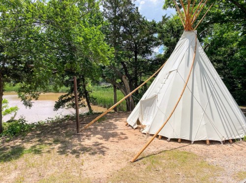 A white teepee stands near a calm river, surrounded by lush green trees and grass under a bright blue sky.