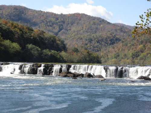 A scenic view of a waterfall cascading over rocks, surrounded by lush green hills and a clear blue sky.