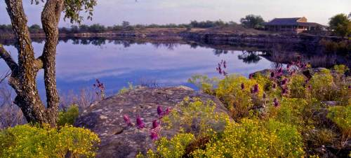 A serene lake surrounded by colorful wildflowers and rocky terrain, with a rustic building in the background.