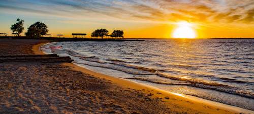 Sunset over a calm beach, with gentle waves lapping at the sandy shore and silhouettes of trees in the background.
