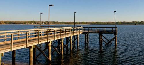 A wooden pier extends over calm water, with lamps lining the walkway and trees in the background under a clear sky.
