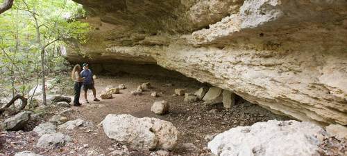 Two people explore a rocky cave with large stone formations and greenery surrounding the entrance.