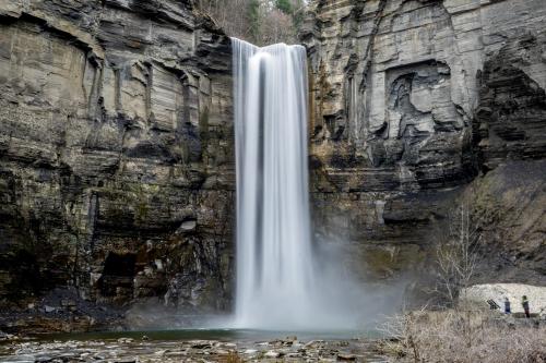 A tall waterfall cascades down rocky cliffs into a serene pool, with two small figures standing nearby.